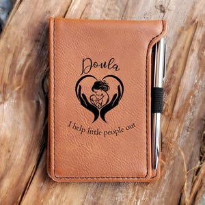 May include: A tan leather-like notebook with a vertical pen holder containing a silver pen. The cover features a black graphic of two hands cradling a baby inside a heart shape, with the text "Doula" and "I help little people out" printed.