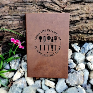 May include: A brown leather-bound journal with a personalized inscription that reads "From the Kitchen of Suzanne - From the Kitchen of". The inscription is surrounded by a circle with a star and a graphic of kitchen utensils.