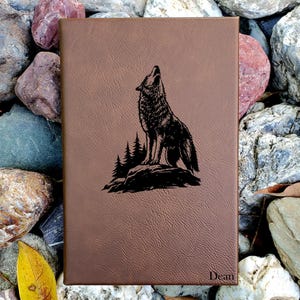May include: A rectangular brown notebook with a textured surface featuring a black engraved illustration of a wolf howling on a rock in front of pine trees. The name Dean is engraved in the bottom right corner of the front cover.
