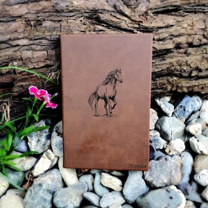 May include: A brown leather journal with a horse engraved on the front. The journal is sitting on a bed of rocks and has the name "Martha" engraved on the bottom.