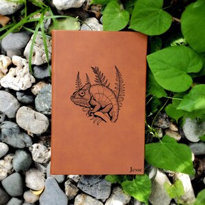 May include: A brown leather journal with a black and white illustration of a chameleon on a branch with leaves. The journal has the name "Jesse" embossed on the bottom right corner.