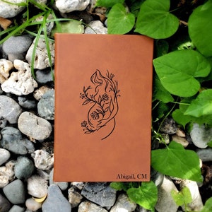 May include: A brown leather journal with a floral design of a pregnant woman. The journal is personalized with the name "Abigail, CM".