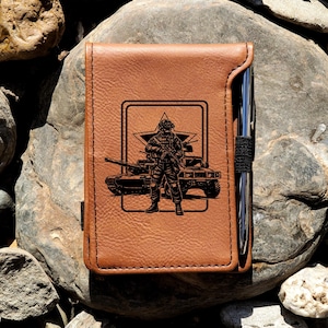 May include: A tan leather notebook cover featuring a black laser-engraved graphic of a soldier standing in front of a military tank and a star emblem. A silver metal pen is held in a black elastic loop attached to the side of the notebook cover.