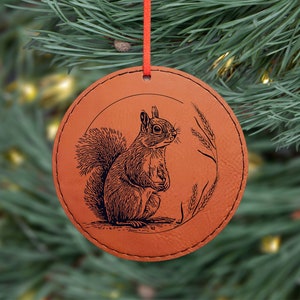 May include: A round, brown leather Christmas ornament with a detailed engraving of a squirrel and wheat stalks. The ornament is suspended by a red cord and is set against a backdrop of green pine needles and soft, out-of-focus lights.