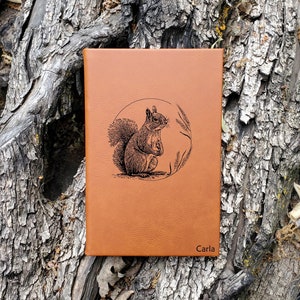 May include: A brown leather journal with a squirrel illustration and the name "Carla" engraved on the front.