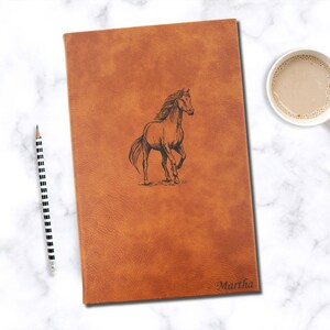 May include: A brown leather journal with a horse design and the name "Martha" engraved on the front.