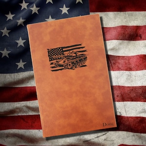 May include: A brown leather journal with a black silhouette of a tank and an American flag on the front. The name "Doug" is engraved on the bottom right corner.