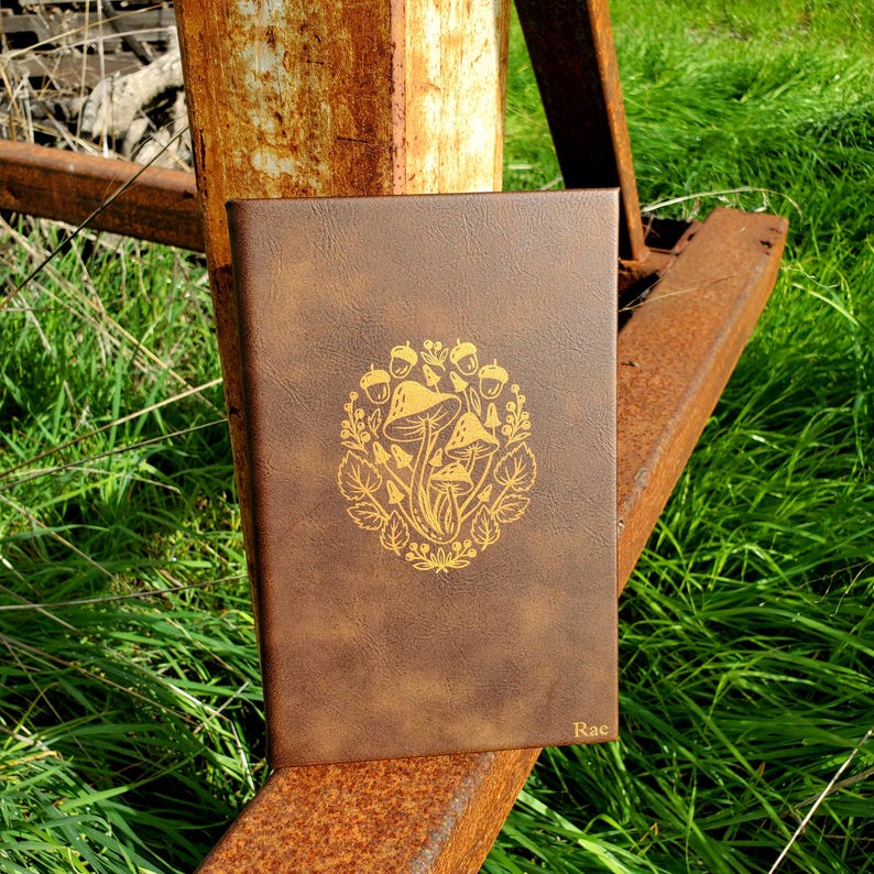 Pu&ograve; includere: Un diario rilegato in pelle marrone con un design in foglia d'oro di funghi e ghiande. Il diario &egrave; personalizzato con il nome "Rae" nell'angolo in basso a destra.