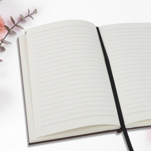 May include: An open notebook with lined pages and a black ribbon bookmark. The notebook has a brown cover and is set against a white background with pink flowers.