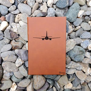 May include: A brown leather journal with a black airplane silhouette on the cover. The name "Doug" is printed in the bottom right corner. The journal is rectangular and appears to be closed.