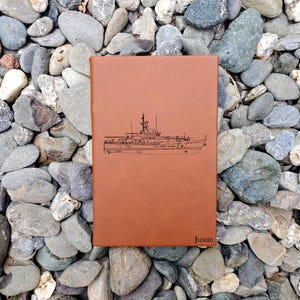 May include: A brown leather journal with a detailed black illustration of a U.S. Coast Guard ship. The journal has the name "Justin" embossed on the bottom right corner. The journal is resting on a bed of gray and tan stones.