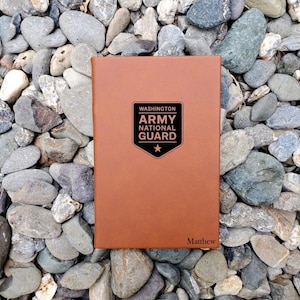 May include: A brown leather journal with the text "WASHINGTON ARMY NATIONAL GUARD" in a black shield design. The name "Matthew" is engraved at the bottom. The journal is resting on a bed of gray and tan stones.