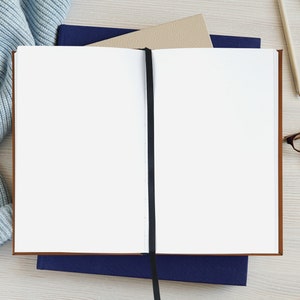 Pu&ograve; includere: Un quaderno bianco vuoto con un segnalibro a nastro nero, aperto su un tavolo di legno. Il quaderno &egrave; circondato da una sciarpa di lana blu, due matite e un paio di occhiali.