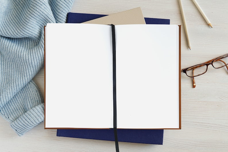 Pu&ograve; includere: Un quaderno bianco vuoto con un segnalibro a nastro nero, aperto su un tavolo di legno. Il quaderno &egrave; circondato da una sciarpa di lana blu, due matite e un paio di occhiali.