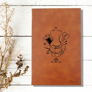 Op de afbeelding: Een bruin lederen notitieboek met een naai-thema ontwerp met een paspop, draad, schaar, spelden en een meetlint. Het notitieboek is gepersonaliseerd met de naam "Karel".