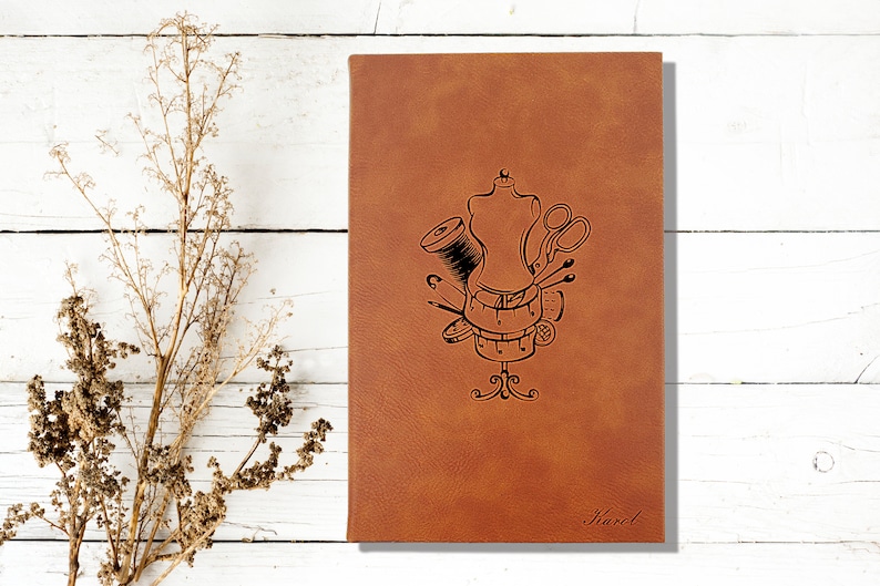 Op de afbeelding: Een bruin lederen notitieboek met een naai-thema ontwerp met een paspop, draad, schaar, spelden en een meetlint. Het notitieboek is gepersonaliseerd met de naam "Karel".