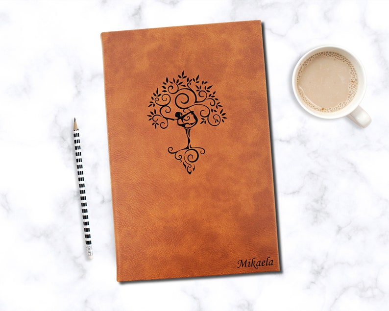 May include: A brown leather journal with a tree of life design and the name Mikaela. A black and white striped pencil and a cup of coffee sit next to the journal on a marble surface.