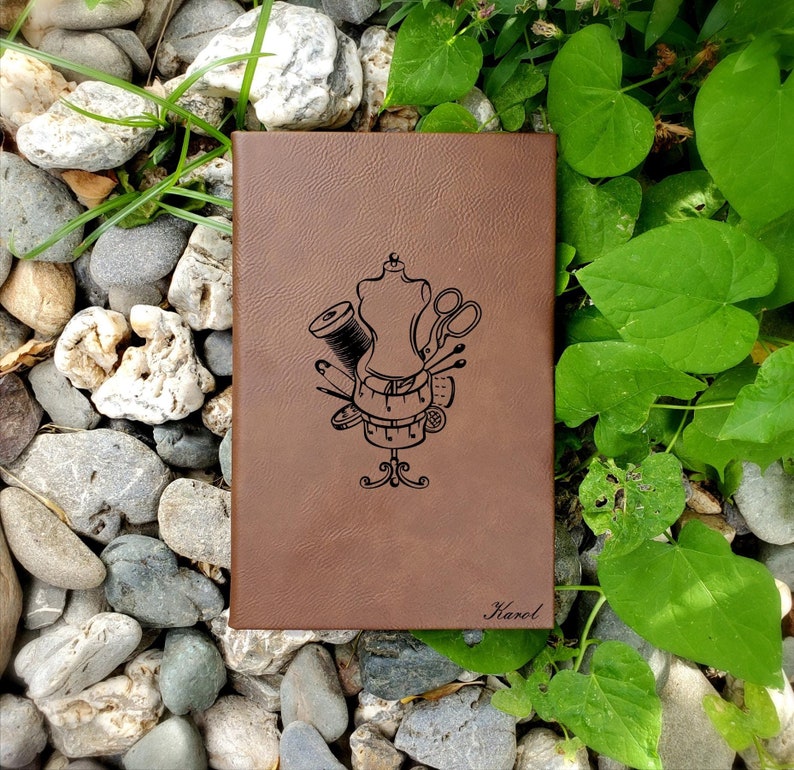 Op de afbeelding: Een bruin lederen dagboek met een ontwerp van een naaipop met gereedschap zoals een schaar, draad en spelden. Het dagboek heeft de naam "Harel" op de voorkant.
