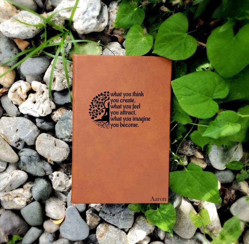 May include: Brown leather journal with a tree of life design and the quote "what you think you create. what you feel you attract, what you imagine you become." The journal is personalized with the name "Aaron".
