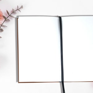 Pu&ograve; includere: Un quaderno bianco vuoto con un segnalibro a nastro nero. Il quaderno &egrave; aperto e disteso su una superficie bianca. Sul lato sinistro dell'immagine ci sono fiori rosa e foglie verdi.