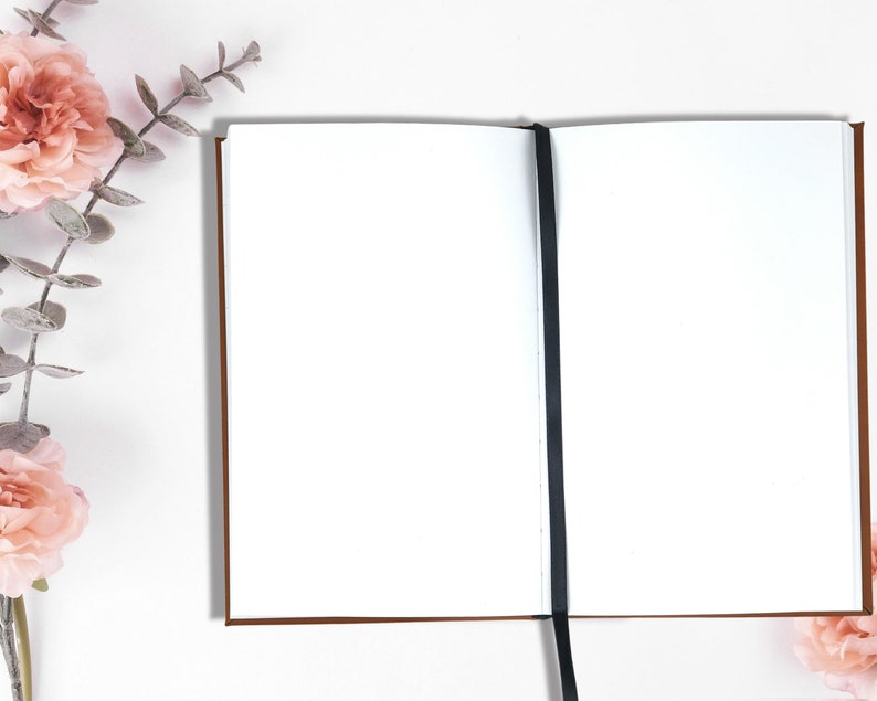 Pu&ograve; includere: Un quaderno bianco vuoto con un segnalibro a nastro nero. Il quaderno &egrave; aperto e disteso su una superficie bianca. Sul lato sinistro dell'immagine ci sono fiori rosa e foglie verdi.
