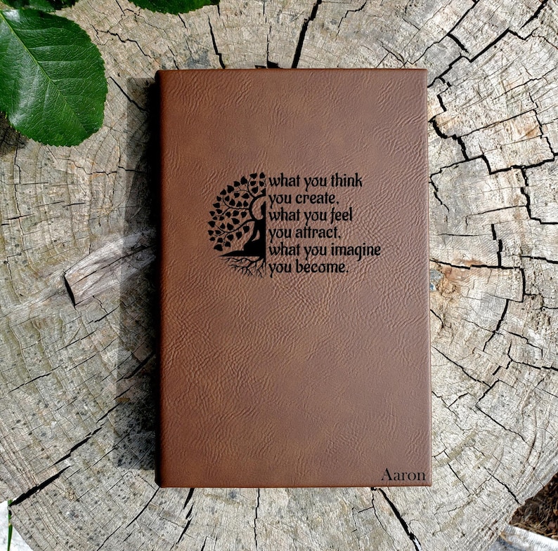 May include: Brown leather journal with a tree of life design and the quote "what you think you create, what you feel you attract, what you imagine you become." The journal is personalized with the name "Aaron".