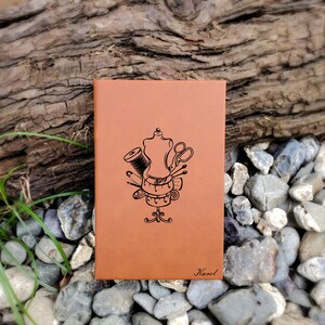 Op de afbeelding: Een bruin lederen notitieboek met een gestikt ontwerp op de voorkant. Het ontwerp bevat een naaivorm, een schaar, draad, naalden en een meetlint. De naam "Karel" is onderaan het notitieboek gegraveerd.