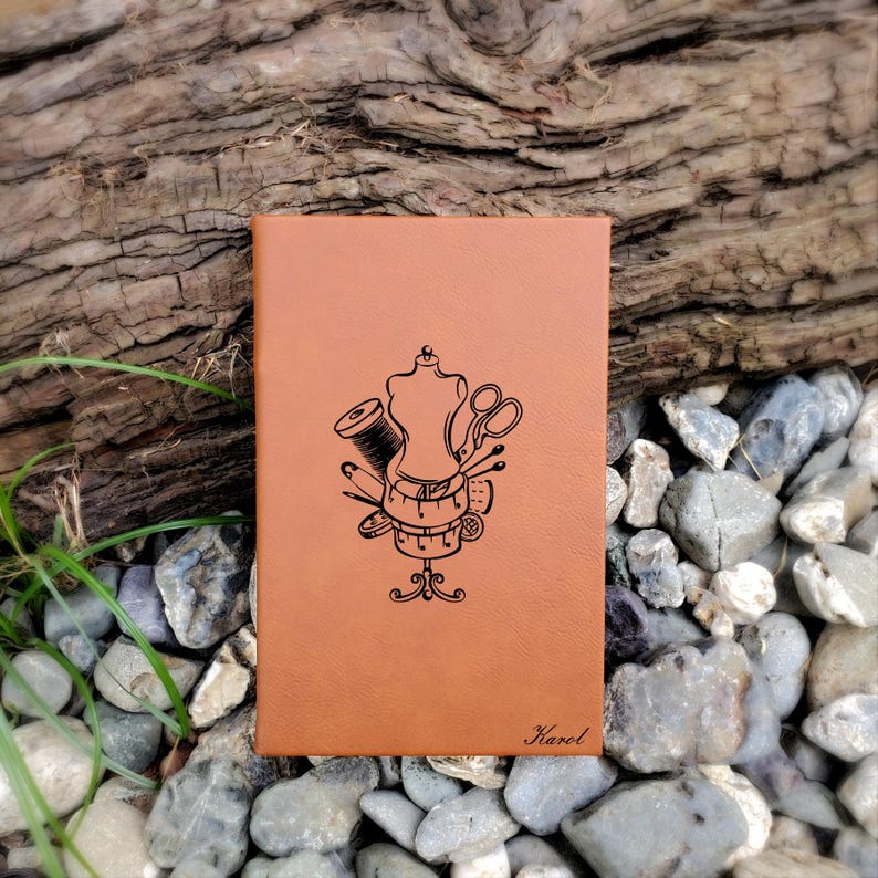 Op de afbeelding: Een bruin lederen notitieboek met een gestikt ontwerp op de voorkant. Het ontwerp bevat een naaivorm, een schaar, draad, naalden en een meetlint. De naam "Karel" is onderaan het notitieboek gegraveerd.