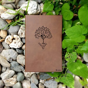 May include: A brown leather journal with a tree design and the name "Jessie" embossed on the cover. The tree design features a person in a yoga pose. The journal is surrounded by rocks and greenery.
