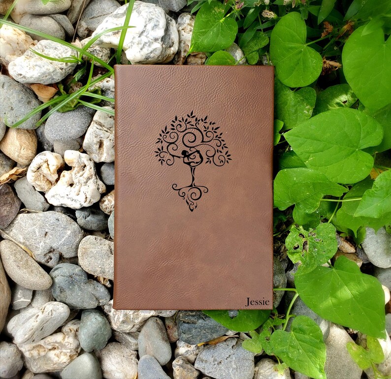 May include: A brown leather journal with a tree design and the name "Jessie" embossed on the cover. The tree design features a person in a yoga pose. The journal is surrounded by rocks and greenery.