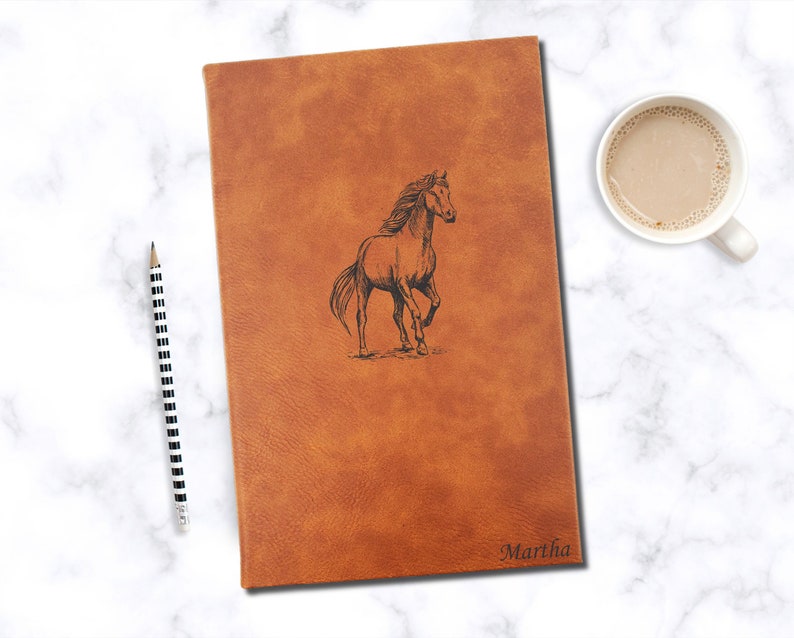 May include: A brown leather journal with a horse design and the name "Martha" engraved on the front.
