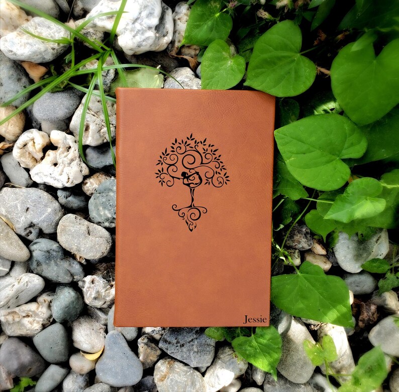 May include: A brown leather journal with a tree of life design and the name "Jessie" embossed on the cover. The journal is surrounded by rocks and green leaves.