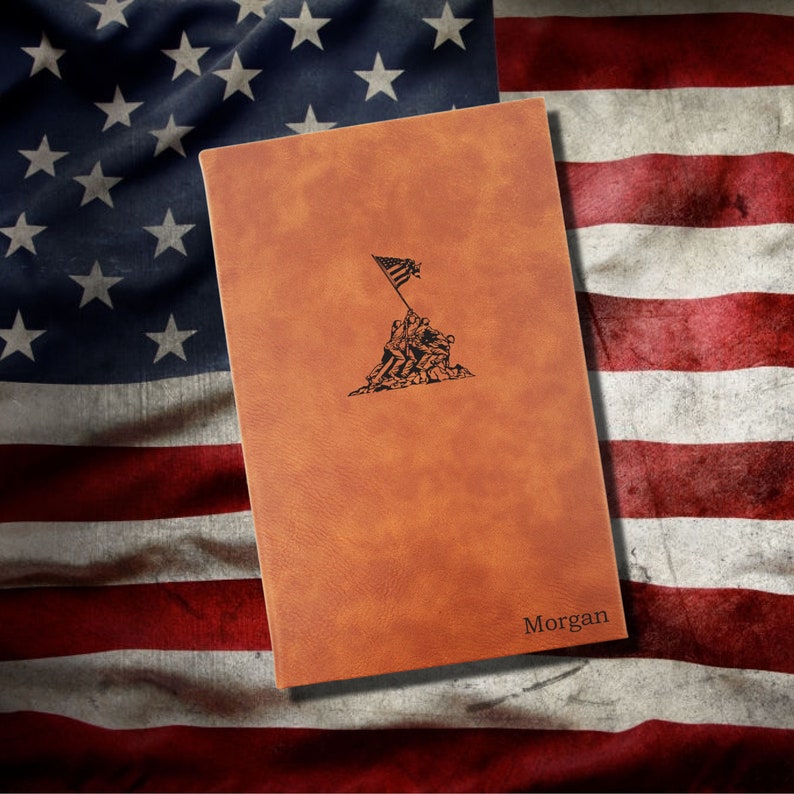 May include: A brown leather journal with a black silhouette of the Iwo Jima flag raising on the front. The journal has the name "Morgan" embossed on the bottom right corner. The journal is sitting on a folded American flag.