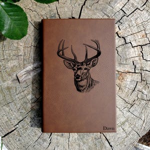 May include: A brown leather journal with a deer head engraved on the front. The journal has the name "Dawn" engraved on the bottom right corner.