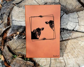 May include: A brown leather journal with a black silhouette of two calves peeking out of a frame. The journal has the name "Isabel" engraved on the bottom.