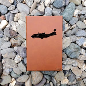 May include: A brown leather journal with a black silhouette of a Coast Guard airplane. The journal has the name "John" embossed at the bottom right corner. The journal is resting on a bed of gray and tan stones.