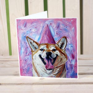 Puede incluir: Una tarjeta de cumpleaños con una pintura de acuarela de un perro Shiba Inu con un gorro de fiesta rosa. El perro está sonriendo con la lengua fuera. El fondo es un diseño abstracto rosa y morado.