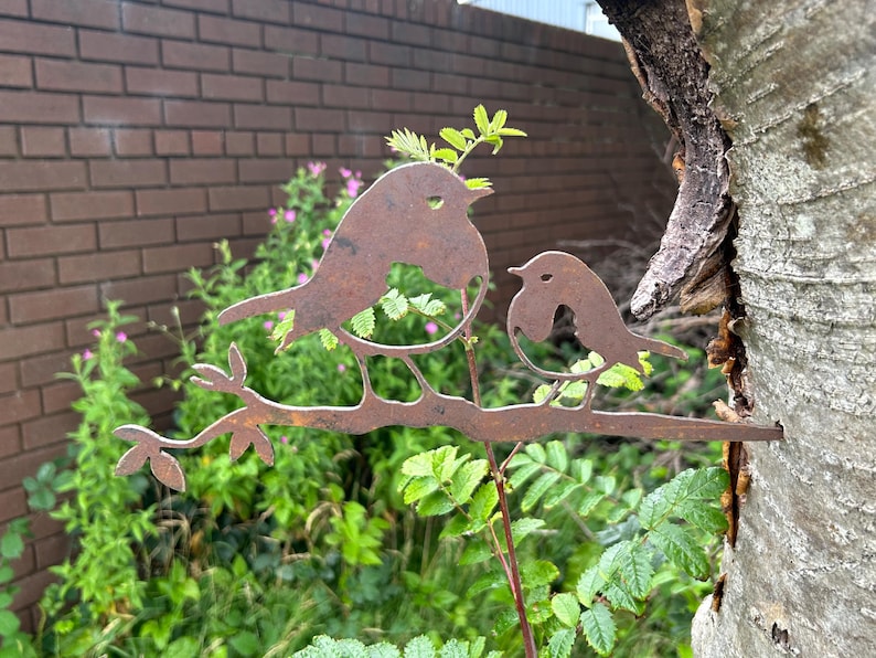 May include: A weathered metal sculpture of two birds on a branch, set against a backdrop of foliage and a brick wall. The birds are depicted in a simple, rustic style, with a natural, aged finish. The sculpture is mounted on a tree.