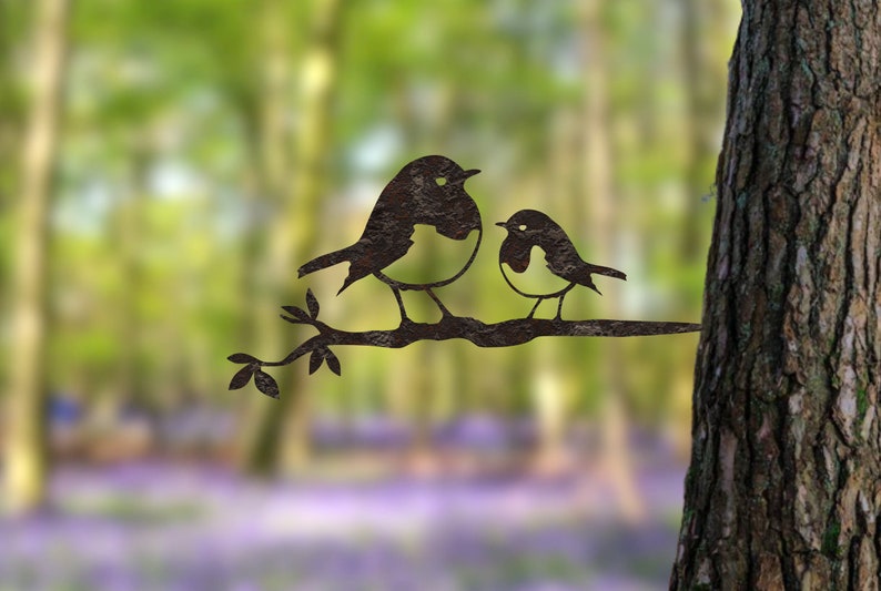 May include: Metal art sculpture of two birds on a branch. The birds are dark with white and black details. The branch has leaves and is set against a blurred forest background. A decorative piece for the garden.