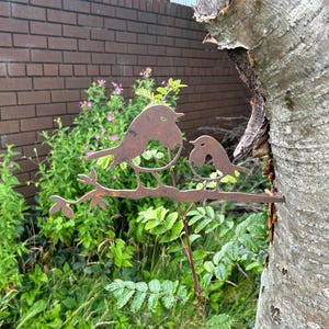 May include: A weathered metal sculpture of two birds on a branch, set against a backdrop of greenery and a brick wall. The birds are facing each other. The sculpture is made of a rusty metal and is attached to a tree trunk.