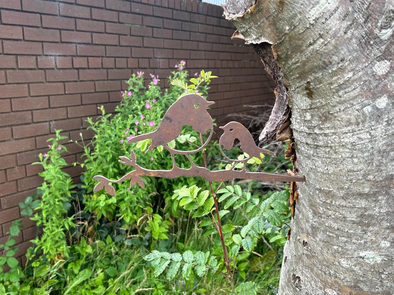 May include: A weathered metal sculpture of two birds on a branch, set against a backdrop of greenery and a brick wall. The birds are facing each other. The sculpture is made of a rusty metal and is attached to a tree trunk.
