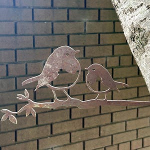 May include: A weathered metal sculpture of two birds on a branch. The birds and branch have a brown finish, set against a brick wall and a tree trunk. The sculpture is designed for outdoor use.