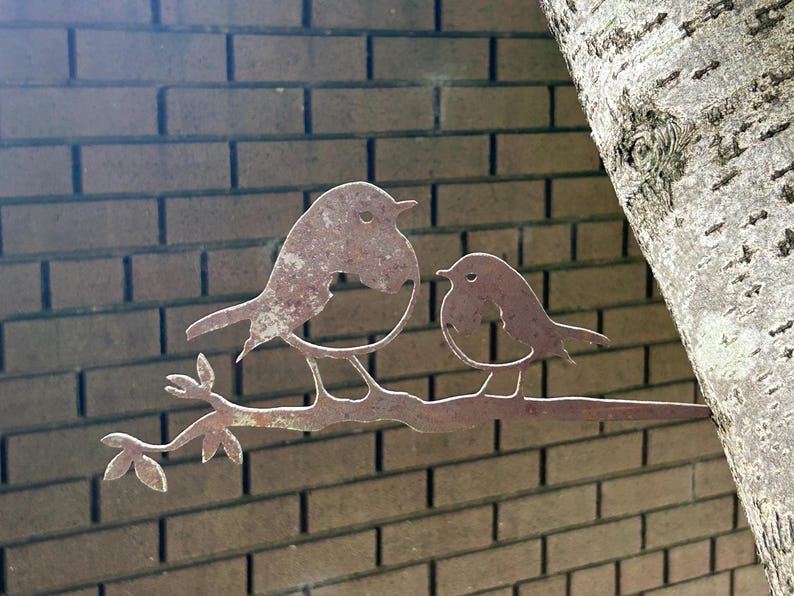 May include: A weathered metal sculpture of two birds on a branch. The birds and branch have a brown finish, set against a brick wall and a tree trunk. The sculpture is designed for outdoor use.