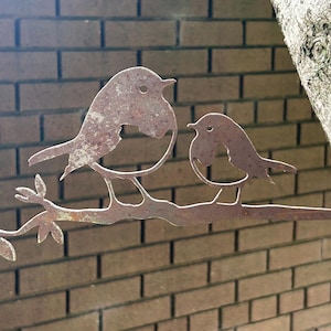 May include: A rusty metal sculpture of two birds perched on a branch with leaves. The birds are stylized with simple shapes and details. The sculpture is mounted against a brick wall and a tree trunk.