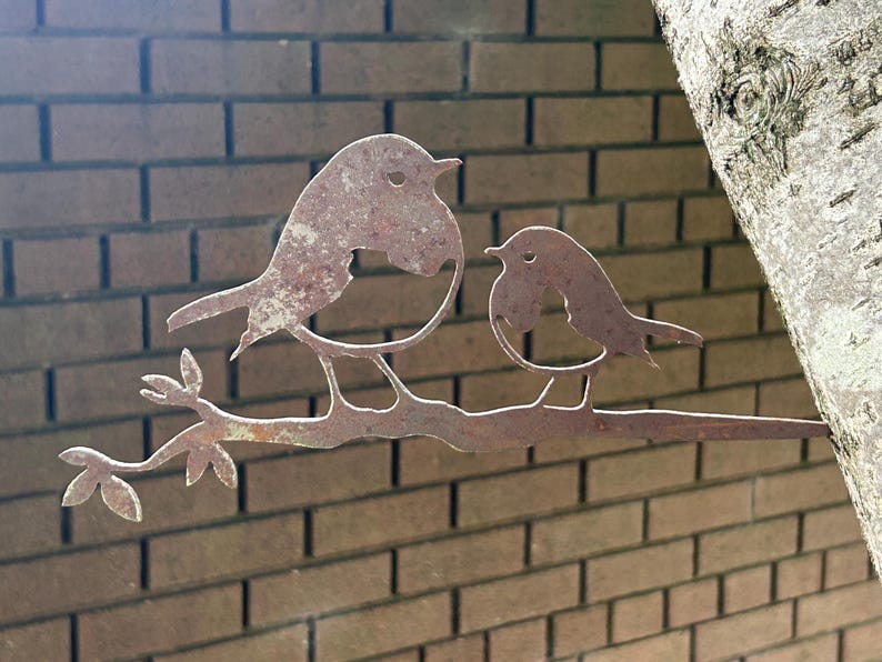 May include: A rusty metal sculpture of two birds perched on a branch with leaves. The birds are stylized with simple shapes and details. The sculpture is mounted against a brick wall and a tree trunk.