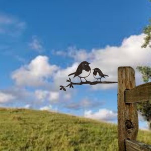 May include: Metal artwork featuring two birds on a branch. The birds are dark brown with white chests, set against a blue sky with white clouds. The artwork is mounted on a wooden fence post, suitable for garden or outdoor decor.