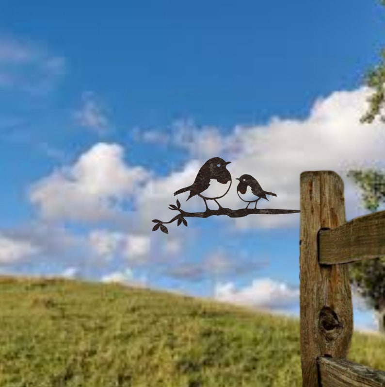 May include: Metal artwork featuring two birds on a branch. The birds are dark brown with white chests, set against a blue sky with white clouds. The artwork is mounted on a wooden fence post, suitable for garden or outdoor decor.