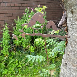 May include: A decorative metal sculpture of two birds on a branch, with a rusty brown finish. The sculpture is set against a backdrop of green plants, a brick wall, and a tree trunk. Suitable for outdoor display in a garden or yard.