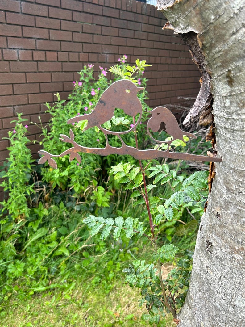 May include: A decorative metal sculpture of two birds on a branch, with a rusty brown finish. The sculpture is set against a backdrop of green plants, a brick wall, and a tree trunk. Suitable for outdoor display in a garden or yard.