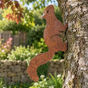 May include: A flat, rust-coloured metal silhouette of a squirrel attached to the side of a tree trunk. The squirrel figure is depicted in a vertical climbing position with a long, curved tail and a small circular cutout near the head area.